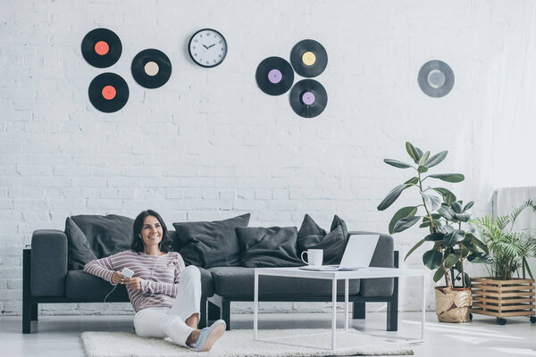 cheerful woman listening music in earphones and holding smartphone while sitting on floor at home