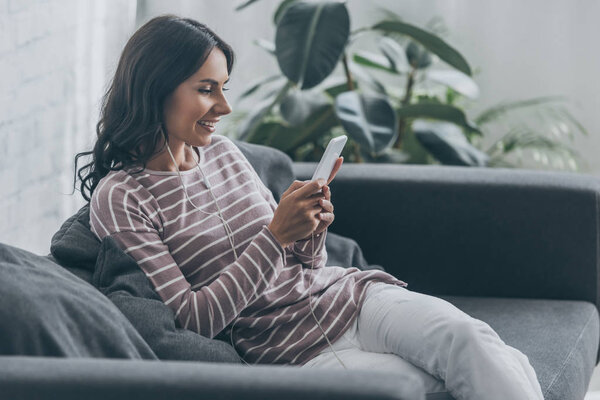 cheerful woman listening music in earphones while sitting on sofa and using smartphone