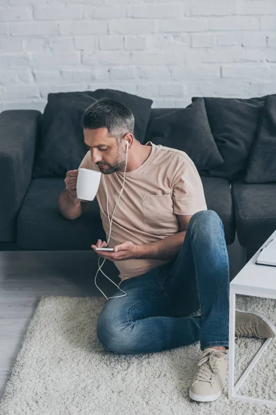 床に座る大人の男性 イヤホンで音楽を聴く スマートフォンを使ってコーヒーを飲む — ストック写真