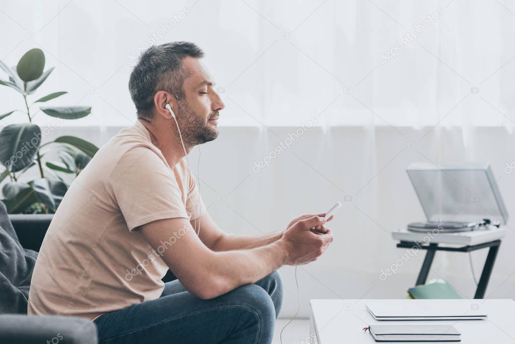 dreamy man in earphones sitting with closed eyes and holding smartphone