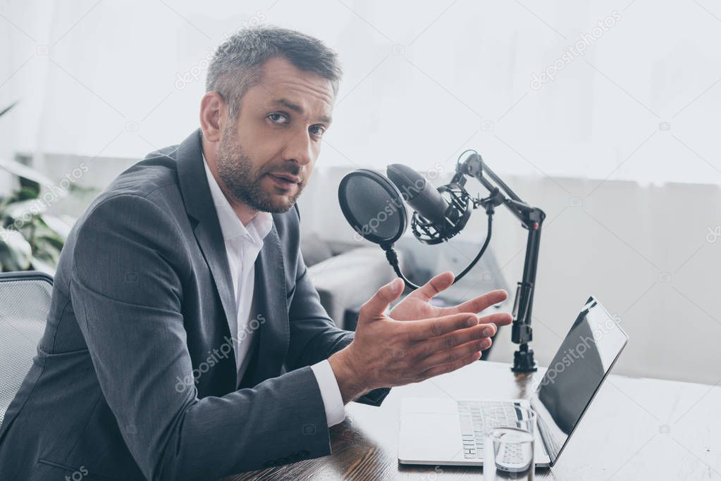 handsome radio host gesturing and looking at camera while sitting at workplace in broadcasting studio