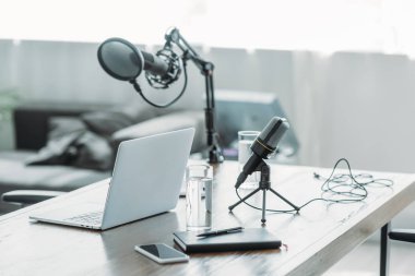 Üzerinde mikrofon, dizüstü bilgisayar, akıllı telefon, defter ve su bardakları olan ahşap masa.