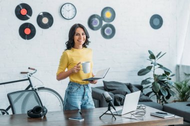 güzel radyo host dizüstü bilgisayar ve kahve fincanı tutarken kameraya gülümseyen