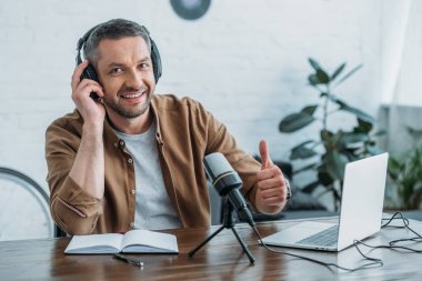 yakışıklı radyo host başparmak gösteren ve işyerinde otururken kamera gülümseyen