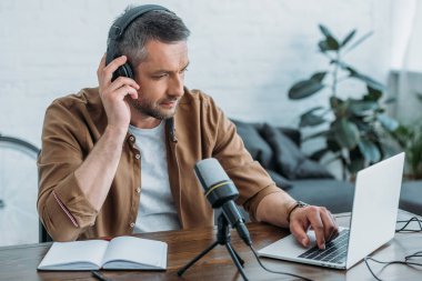 attentive radio host in headphones using laptop while sitting near microphone clipart