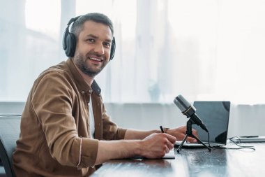 cheerful radio host in headphones smiling at camera while sitting near microphone and laptop clipart