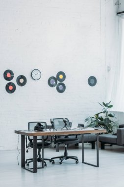 ahşap masa, ofis sandalyeleri ve beyaz duvar üzerinde vinil diskler ile geniş, mobilyalı radyo stüdyosu
