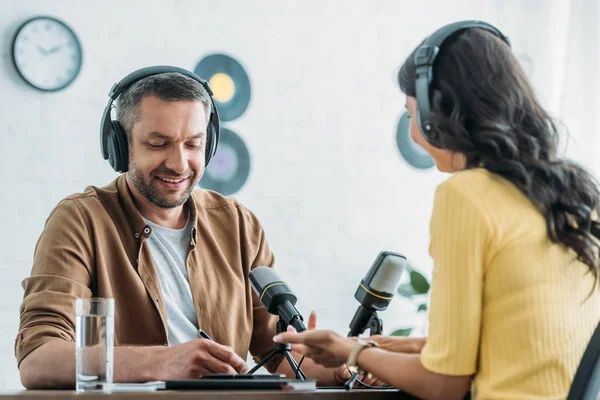 Cheerful Radio Host Headphones Writing Notebook While Sitting Workplace Colleague — Stock Photo, Image