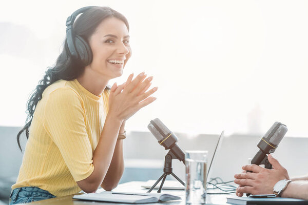 cheerful radio host looking at camera while sitting near colleague in broadcasting studio