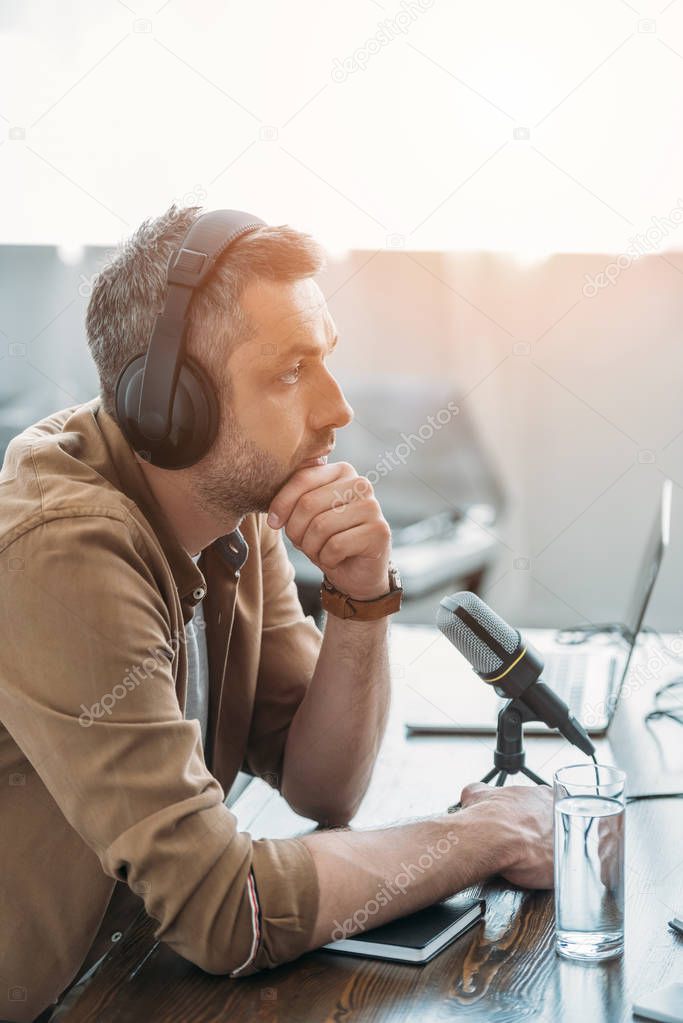 handsome serious radio host in headphones sitting near microphone in broadcasting studio