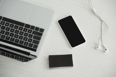 dizüstü bilgisayar üst görünümü, boş ekranlı akıllı telefon, kulaklıklar ve ofis masasında siyah boş kartvizit yığını