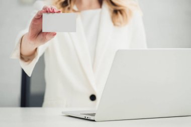 cropped view of businesswoman showing at camera white blank business card while sitting near laptop clipart