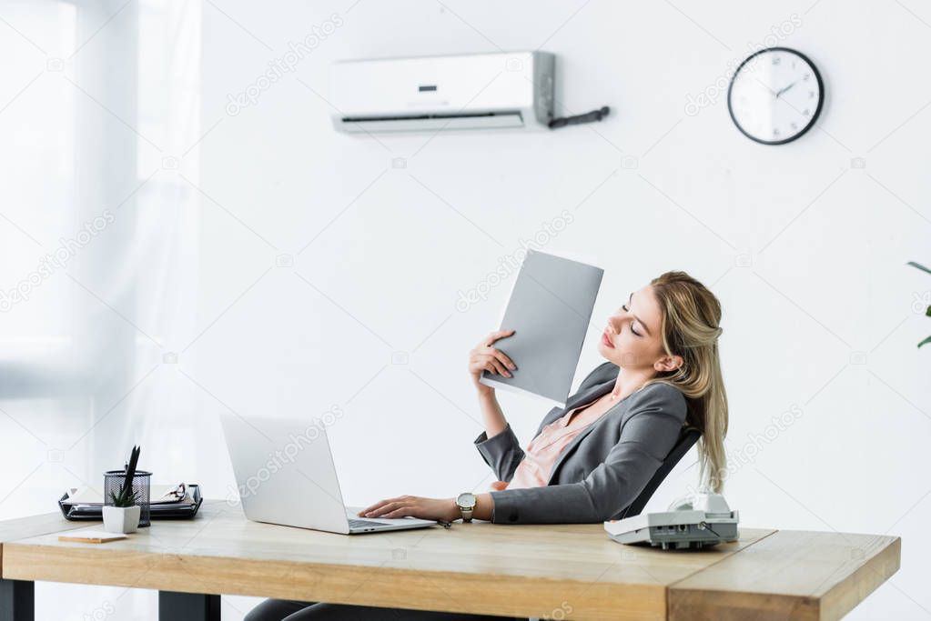 businesswoman sitting in office, holding folder in hand and suffering from heat