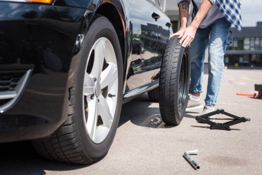 cropped view of man rolling new tire and fixing broken auto, car insurance concept clipart