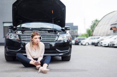upset businesswoman sitting near broken auto with smartphone in hands, car insurance concept clipart