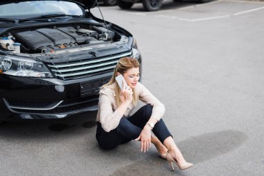 sad businesswoman talking on smartphone while sitting near broken auto, car insurance concept clipart