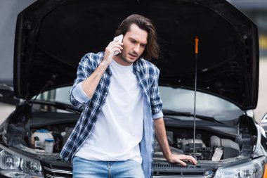 frustrated man talking on smartphone while standing near broken auto with open trunk, car insurance concept clipart