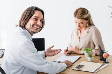 selective focus of happy doctor looking at camera while patient signing document clipart