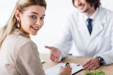 selective focus of patient sitting with doctor behind wooden table, signing document and looking at camera clipart