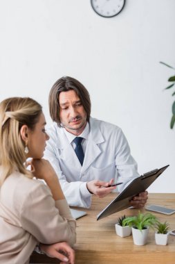 yakışıklı doktor elinde belge tutan, anlaşma kalem ile işaret ve hastanın yanında ahşap masanın arkasında oturan