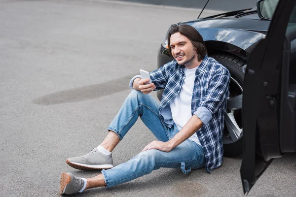 スマートフォンを手に壊れた自動車の近くに座るカジュアルな服を着た幸せな男 自動車保険コンセプト — ストック写真