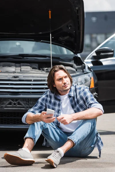 スマートフォンを手に持ったハンサムな男 目を離して見て 開いたトランクで壊れた自動車の近くに座って 自動車保険の概念 — ストック写真