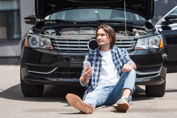男子在休闲服装手拿着智能手机 看着远方 坐在附近的破碎的汽车与开放的后备箱 汽车保险的概念 — 图库照片