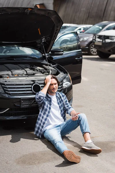 開いたトランクで壊れた自動車の近くに座って 頭に手を握っているスマートフォンを持つ男 自動車保険の概念 — ストック写真