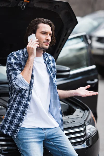 upset man in casual wear talking on smartphone while standing near broken auto with open trunk, car insurance concept