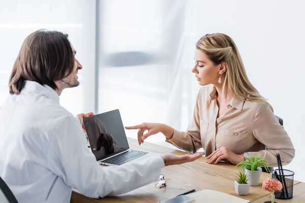 patient sitting with doctor behind wooden table and pointing with finger at laptop display