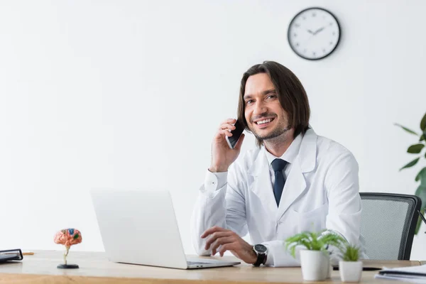 Yakışıklı Doktor Akıllı Telefon Konuşuyor Kamera Bakıyor Masada Dizüstü Bilgisayar — Stok fotoğraf