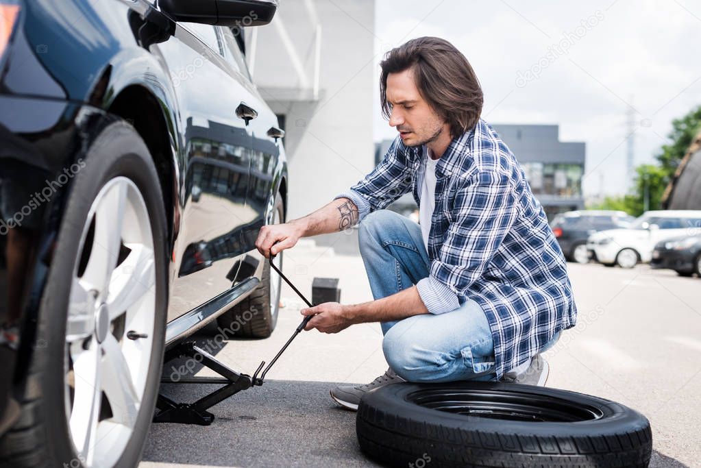 man in casual wear using jack tool and changing broken wheel on auto, car insurance concept