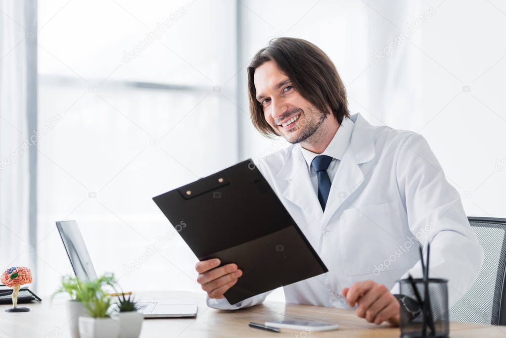 happy doctor with clipboard in hand looking at camera while sitting in office