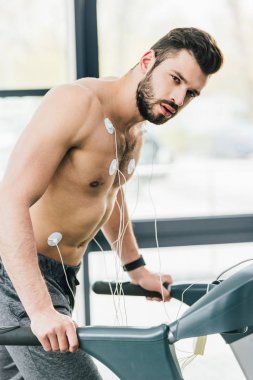 spor salonunda dayanıklılık testi sırasında koşu bandı üzerinde çalışan elektrotlar ile yakışıklı sporcu ve kamera bakarak