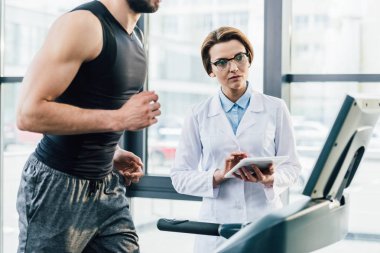 spor salonunda dayanıklılık testi sırasında Dijital Tablet ile doktor yakınında koşu bandı üzerinde çalışan sporcu