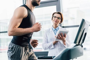 spor salonunda dayanıklılık testi sırasında Dijital Tablet ile doktor yakınında koşu bandı üzerinde çalışan sporcu
