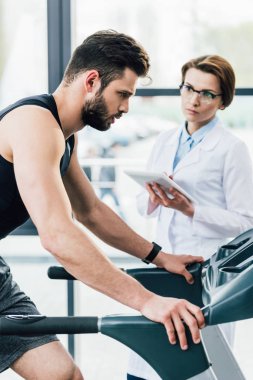 yakışıklı sporcu spor salonunda dayanıklılık testi sırasında doktor yakınında koşu bandı üzerinde çalışan