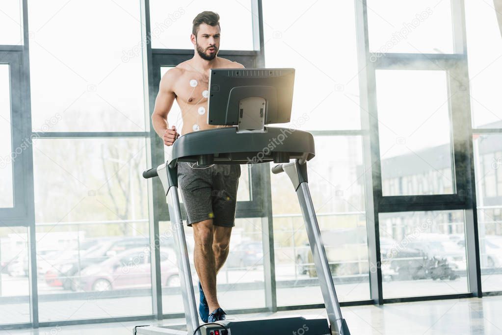 handsome sportsman with electrodes running on treadmill during endurance test in gym with daylight