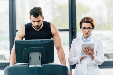 yakışıklı sporcu spor salonunda dayanıklılık testi sırasında doktor yakınında koşu bandı üzerinde çalışan