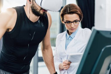 spor merkezinde dayanıklılık testi sırasında Dijital Tablet ile doktor yakınında koşu bandı üzerinde çalışan sporcu