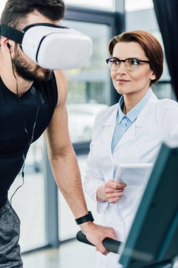 spor merkezinde dayanıklılık testi sırasında Dijital Tablet ile doktor yakınında koşu bandı üzerinde çalışan sporcu