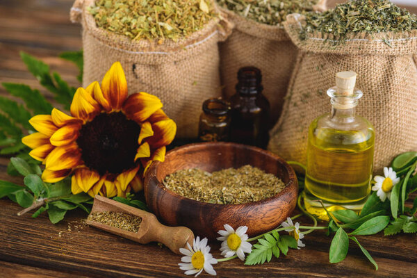 wooden bowl, spatula, sackcloth bags with dried herbs, bottles with essential oils, sunflower and chamomile flowers on wooden surface 