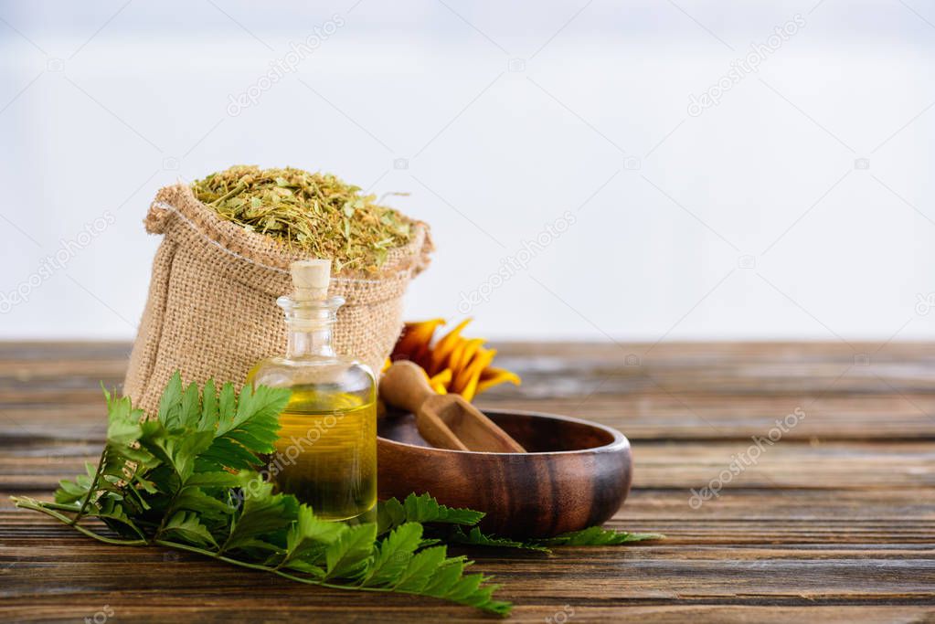 sackcloth bag with dried herbs, corked bottle with essential oil, fern leaves, wooden bowl and spatula on wooden surface