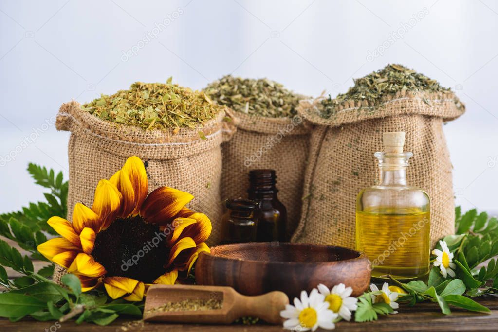sackcloth bags with dried herbs, bottles with essential oils, sunflower and chamomile flowers, wooden bowl and spatula on wooden surface