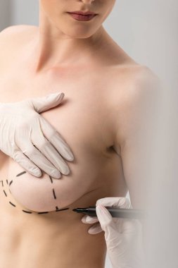 cropped view of plastic surgeon in latex gloves making marks on breast isolated on grey clipart