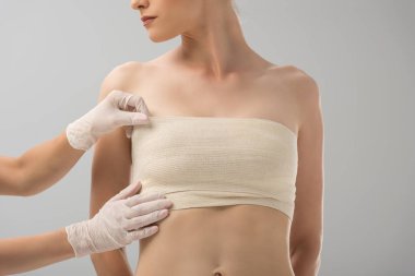 lateks eldivenlerde plastik cerrahın kısmi görünümü ve gri üzerinde izole edilmiş meme bandajında hasta