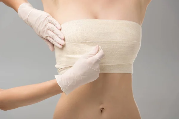 乳胶手套中整形外科医生的部分视图和在灰色上隔离的乳房绷带患者 — 图库照片