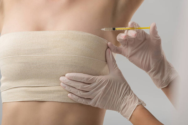 partial view of plastic surgeon in latex gloves doing injection and patient in breast bandage isolated on grey