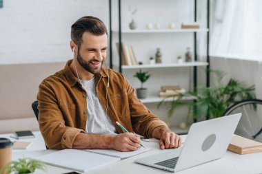 good-looking man listening music, using laptop and writing in notebook  clipart