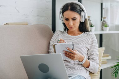 beautiful woman with headphones writing in notebook and holding laptop  clipart